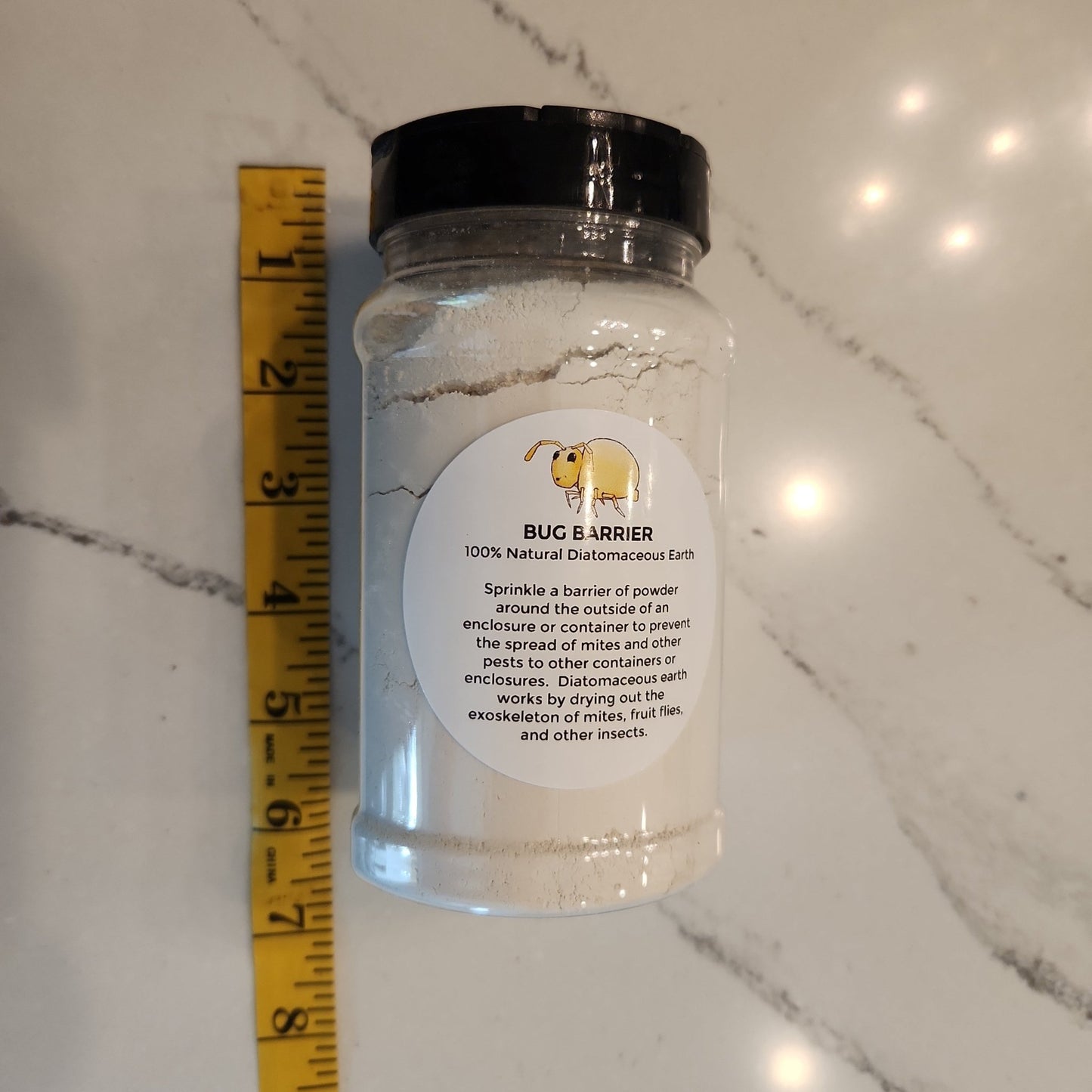 A clear plastic jar with a black lid, labeled Diatomaceous Earth Mite/Bug Barrier by I Heart Bugs, contains white powder and sits on marble near a yellow measuring tape. The label shows a cartoon dog and mite prevention instructions.