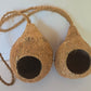 The Hanging Coconut Hideout by IHEARTBUGS, INC. features two hollowed coconut shells joined by a thick, braided rope, forming an inviting hideaway. Each shell displays a wide circular opening facing forward, with their rough, fibrous surfaces and natural brown tones standing out against a plain, light background.