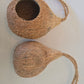Two brown Hanging Coconut Hideouts from IHEARTBUGS, INC.—each featuring natural fibers and a twisted rope handle—rest on a white surface; one hideout is whole, the other is halved with an open top, making them perfect as natural shelters or cozy hideouts for small pets.