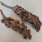 Two brown, woody stems—sold as Palm Blossom Stalk by I Heart Bugs—are covered in layers of curled, overlapping, rough-edged, dark brown mushroom caps and displayed on a plain white background. The mushrooms have a dry, textured appearance with irregular shapes and varying colors throughout.