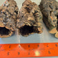 Three hollow, rough Small Cork Bark Rounds by IHEARTBUGS, INC. are placed side by side on a light blue surface. An orange ruler shows they measure about 10–12 cm long—ideal for use in bioactive enclosures.