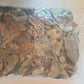A clear plastic bag of I Heart Bugs Tropical Leaf Litter Mix, containing organic leaves in natural shades of brown, yellow, and green, lies flat on a light-colored surface. The assorted crinkled leaves vary in size and shape, making them ideal as an add-on for live animal purchases for use in bioactive enclosures or blackwater aquariums.